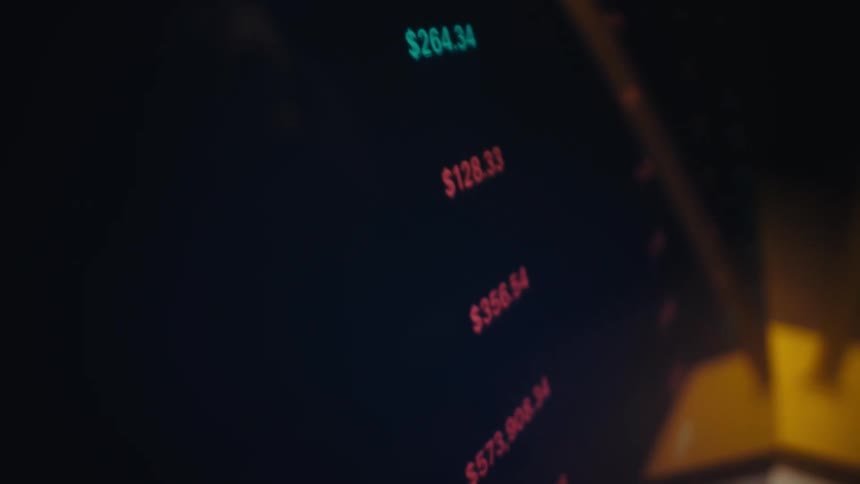 A close up shot of a dark digital screen displays a list of cryptocurrency data and financial charts with shifting green and red lines