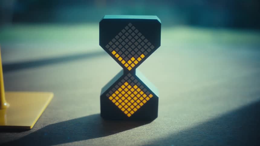 A black digital hourglass sits on a flat surface in sunlight featuring a grid of led pixels that animate downward from the top triangular section to the bottom mimicking the flow of sand in a traditional timer as the light patterns shift