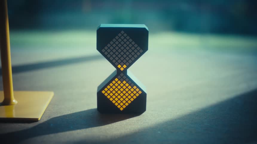 A black digital hourglass sits on a table while yellow led pixels fall from the top section to the bottom mimicking sand and a yellow stand is visible to the left against a blurred outdoor background as sunlight hits the surface
