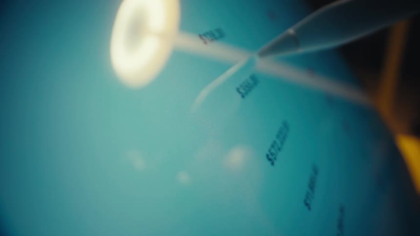 A person uses a white stylus to scroll through a cryptocurrency market list on a tablet displaying various coin logos and financial values while a ring light reflects on the glass surface and the camera pans across the digital interface
