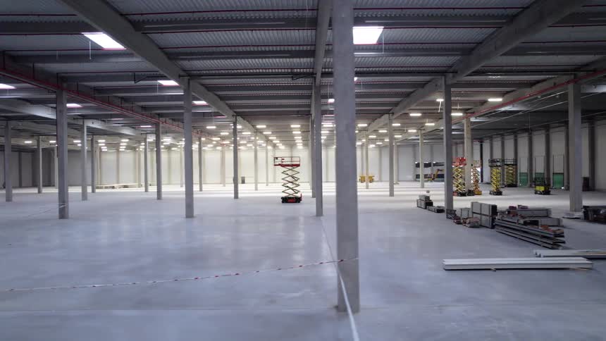 An empty largescale industrial warehouse under construction featuring high ceilings concrete pillars and various construction equipment like scissor lifts and metal materials scattered across a polished floor under bright overhead lighting