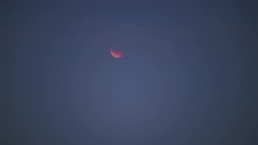 Reddish moon partially obscured in a uniform blue sky