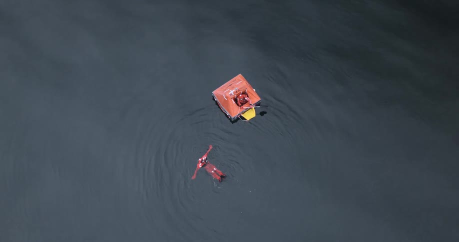 Person wearing rescue gear floating in the water near an orange inflatable rescue raft