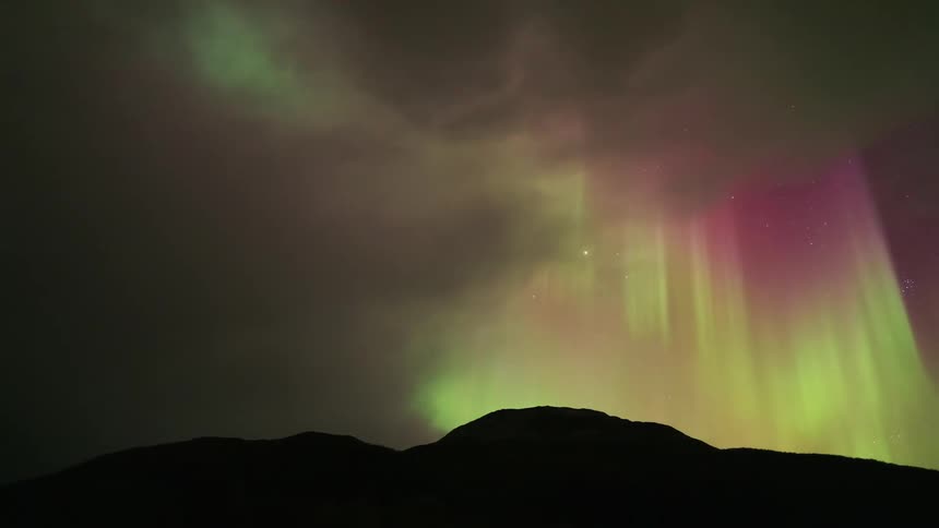 Following the mesmerizing dance of the green and pink aurora borealis as they shimmer across the starry night sky above a dark mountain silhouette