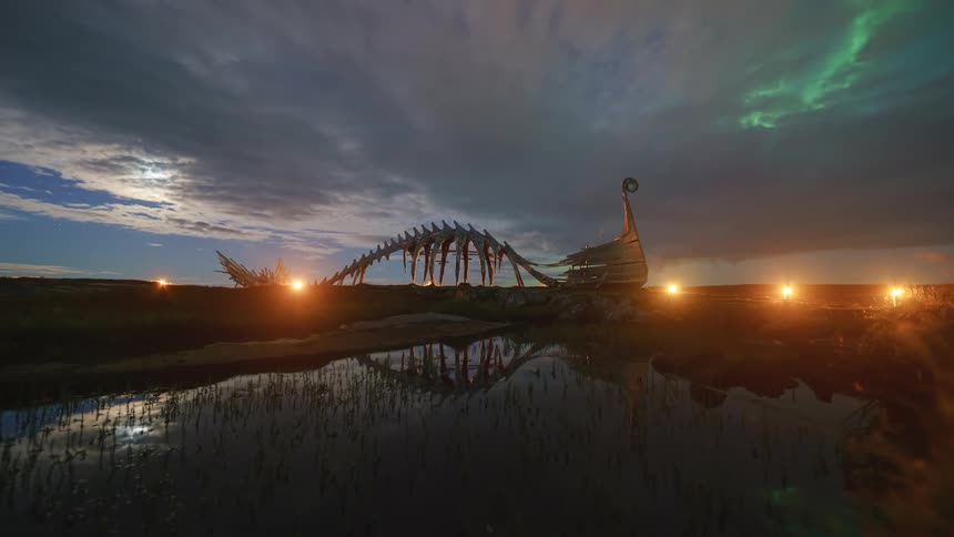 A viking ship skeleton stands silhouetted against a dramatic twilight sky with faint northern lights and reflections shimmering in the water below