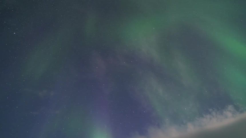 The aurora borealis dances across a dark starry sky in vibrant green hues shifting and shimmering above a thin layer of white clouds that move slowly along the bottom of the frame during a clear night
