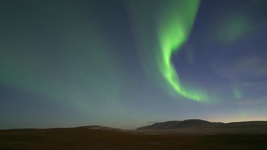 A time lapse captures the vibrant green northern lights dancing and swirling across a star filled night sky over a dark vast landscape of hills and plains