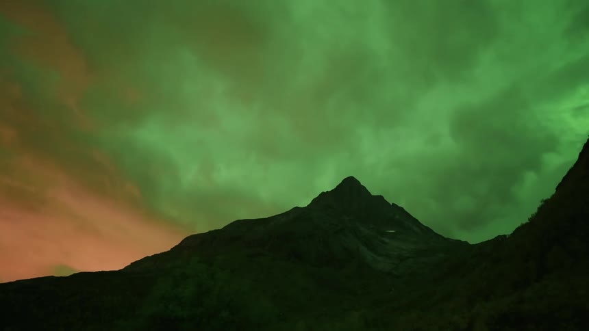 A timelapse captures vibrant green northern lights shimmering and shifting across a dark cloudy sky above the silhouette of a sharp mountain peak