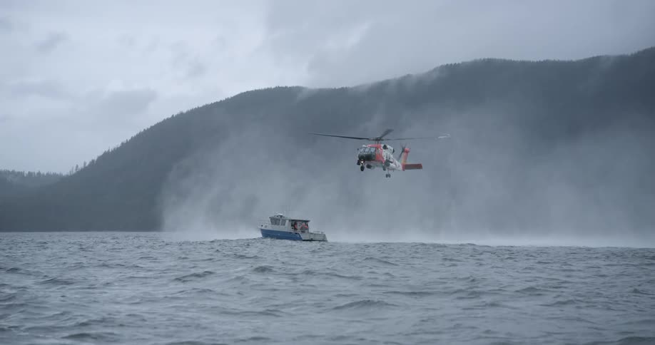 Helicopter hovering over a vessel in rough sea near a mountainous coastline under mist