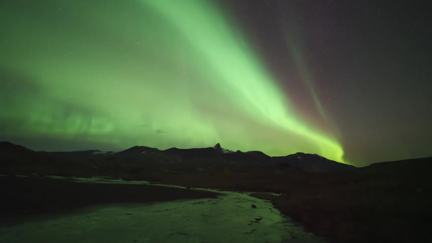 Brilliant green and purple ribbons of the aurora borealis dance across the starry night sky illuminating a vast frozen landscape and the dark mountains beyond
