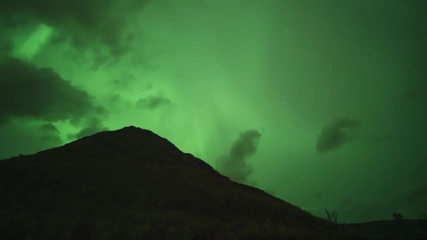 A timelapse video shows the green aurora borealis shimmering and dancing across a dark night sky filled with moving clouds behind a large dark mountain silhouette