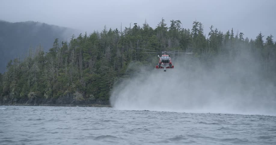 A rescue helicopter hovers low over choppy water near a forested coastline kicking up spray as mountains fade into the mist behind it