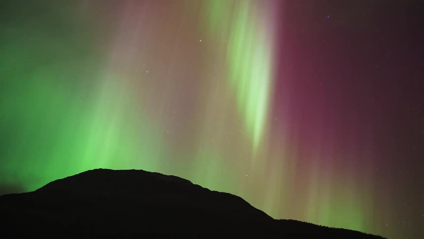 In a stunning timelapse vibrant bands of green and red aurora borealis dance across the starry night sky above a silhouetted mountain