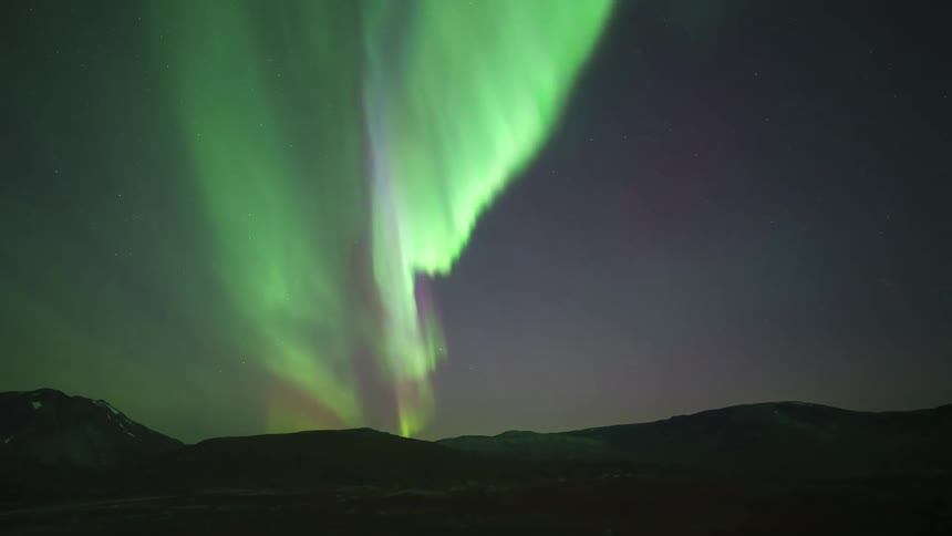 The glowing lights of the aurora borealis move over mountains in the night sky