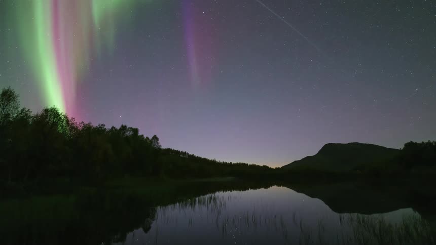Green and purple northern lights stretching across the night sky reflected in a lake surrounded by trees and mountains