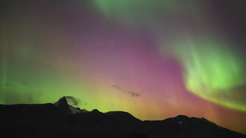 A timelapse captures vibrant green and pink northern lights shimmering and shifting across a dark sky filled with moving clouds above a dark mountain range with a prominent sharp peak