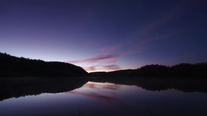 A lake lies still surrounded by dense trees as sunset fades into night the sky above begins to darken and a soft green light hovers overhead casting a surreal glow across the waters surface and the silhouetted treetops