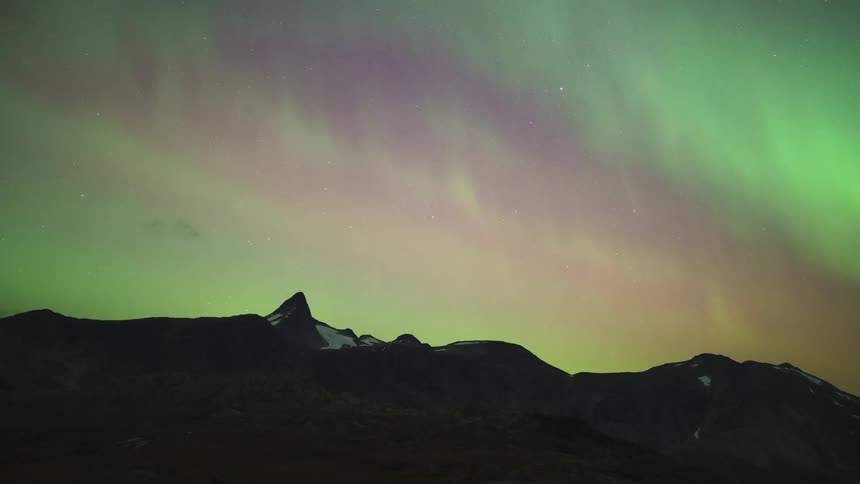 A timelapse captures vibrant green and purple northern lights swirling and dancing across a star filled night sky above a dark mountain range with a sharp peak