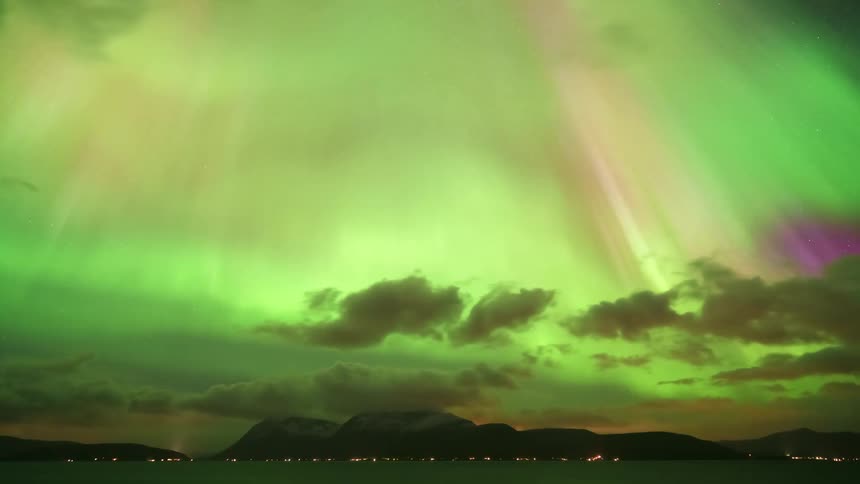 A timelapse captures vibrant green and pink northern lights shimmering and shifting across a dark sky filled with moving clouds above a distant mountain range and a body of water lined with small city lights