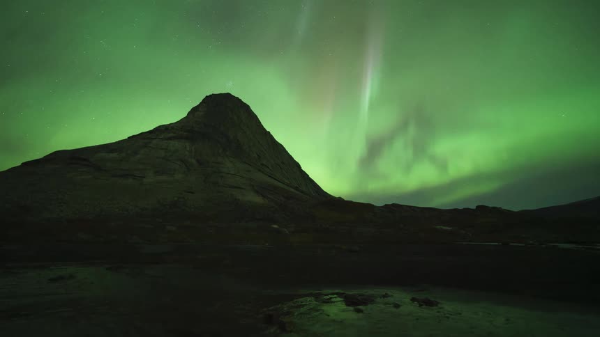 A timelapse captures vibrant green northern lights swirling and dancing across a star filled night sky above a dark rocky mountain peak and a small body of water in the foreground