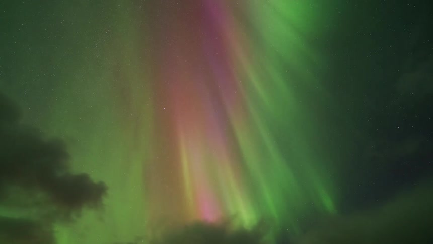 Vibrant green and pink aurora borealis pillars dance across a dark starry night sky above a layer of moving clouds creating a colorful natural light display in the upper atmosphere