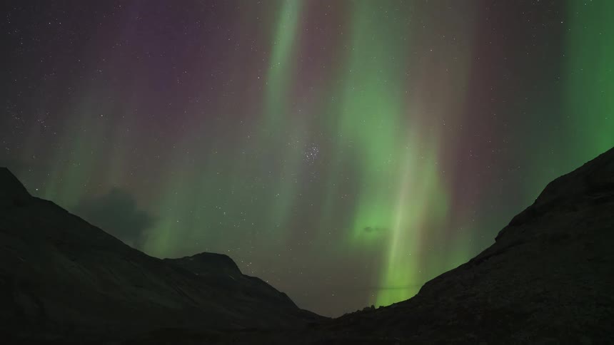 A starfilled night sky stretches over the mountains and a green light hovers above the slopes casting a faint glow across the rugged terrain