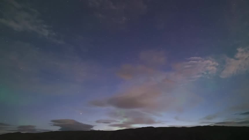 A timelapse captures vibrant green northern lights shimmering and shifting across a star filled night sky above the dark silhouette of a low mountain range