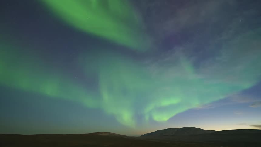 A timelapse captures vibrant green northern lights swirling and dancing across the sky above a dark vast landscape of hills and mountains