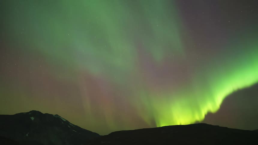 A starfilled night sky stretches over the mountains and a green light hovers above the slopes casting a faint glow across the rugged terrain