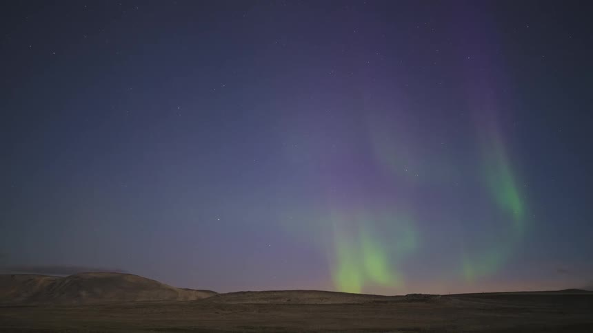 A timelapse captures vibrant green and purple northern lights swirling and dancing across a star filled night sky above a dark landscape of low hills