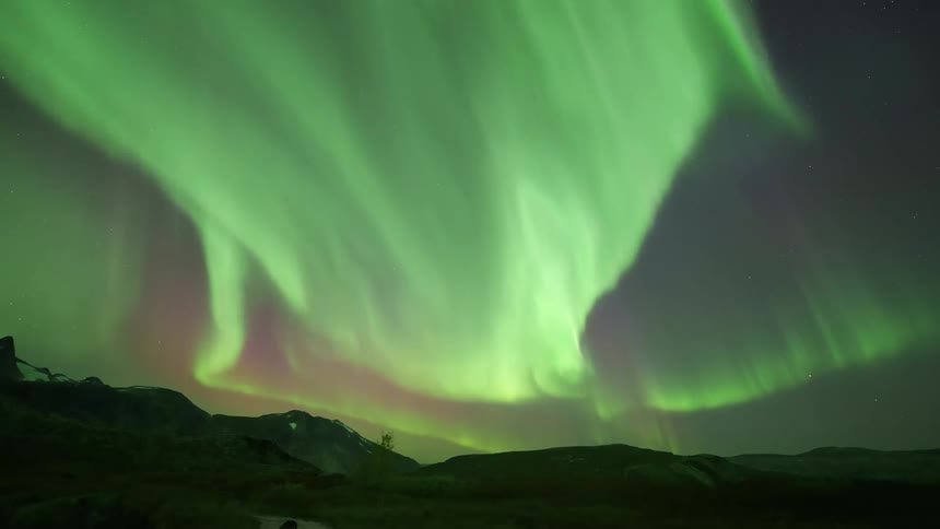 A timelapse captures vibrant green and purple northern lights swirling and dancing across a starfilled night sky while reflecting in a small stream that flows through a dark mountain landscape