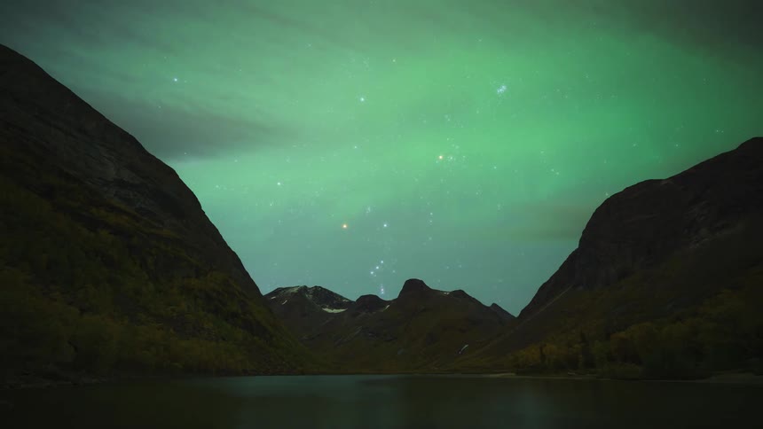 A timelapse captures vibrant green northern lights shimmering and shifting across a star filled night sky above the dark silhouettes of a vast mountain valley and a calm lake