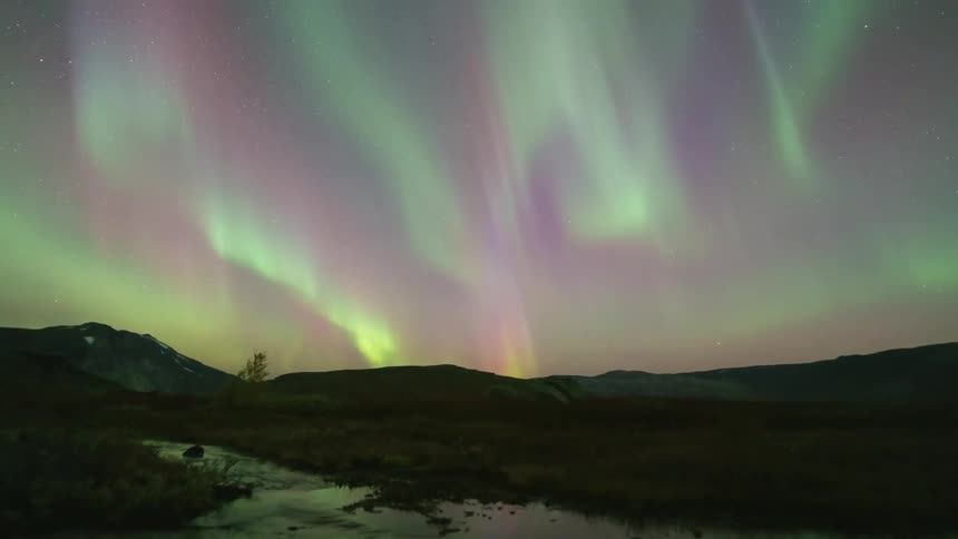 Green and purple aurora borealis dance across a starry night sky above a dark mountain range while a small stream flows through the rocky foreground reflecting the vibrant celestial colors