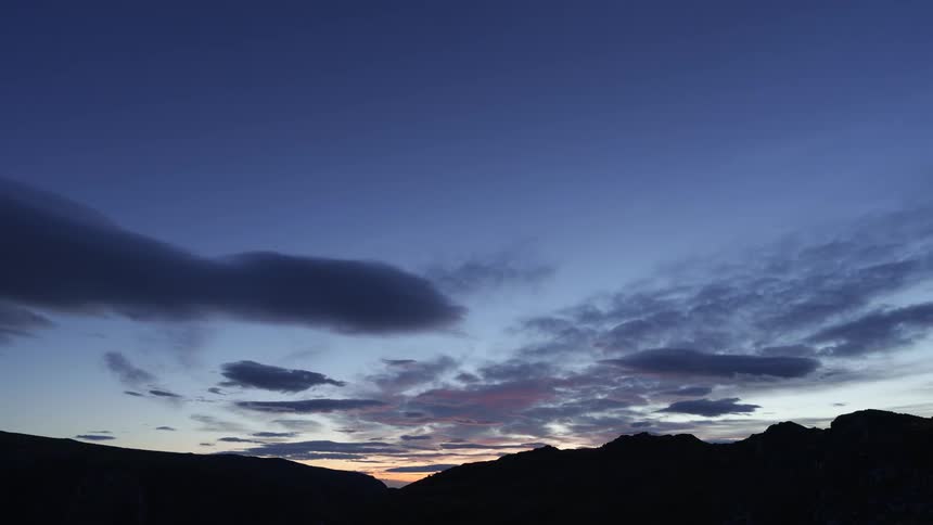 A timelapse captures the transition from a starry night with faint green aurora over silhouetted mountains to a bright dawn as thick layers of clouds move rapidly across the sky eventually obscuring the stars and filling the frame