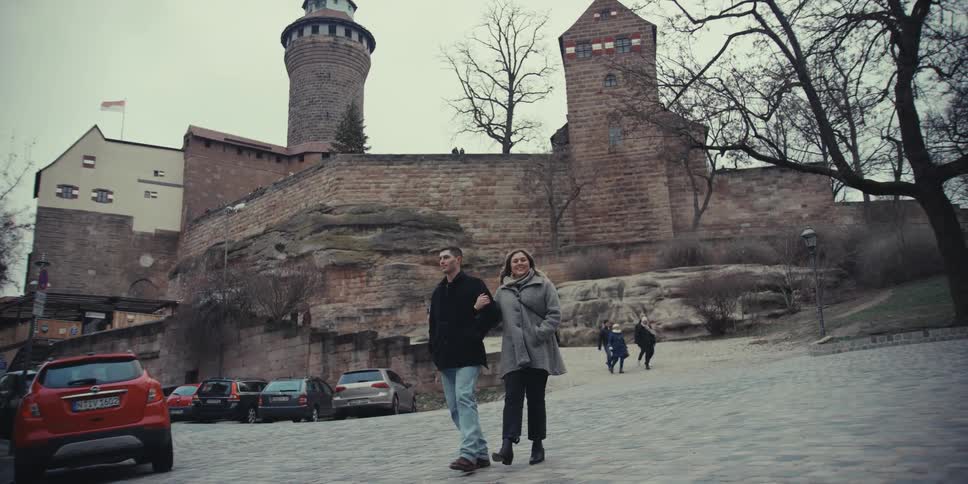 People walk in front of a castle with cars parked on the side of the road
