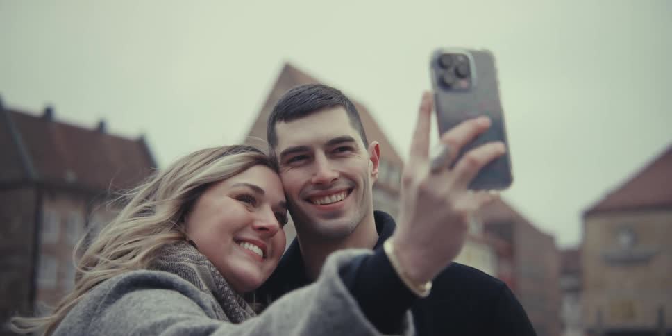 A smiling man and woman stand in a european plaza taking selfies on a phone