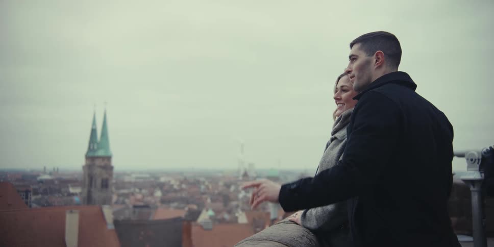 A couple stands side by side pointing fingers overlooking a historic cityscape quietly taking in the panoramic view beneath an overcast sky