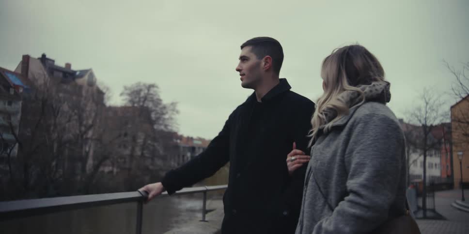 A young man and woman walk arminarm along a riverside path in a european city sharing a laugh before stopping at a railing to look out over the water and distant historic architecture under a cloudy sky