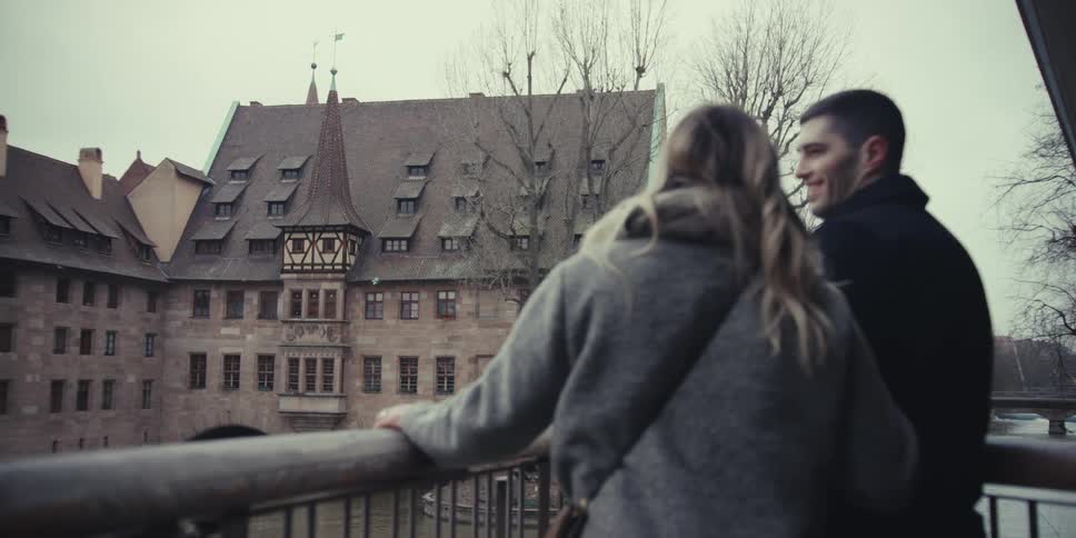 A couple stands on a bridge overlooking a historic building and a river on a cloudy day the man smiles and shares a brief affectionate moment with the woman while they admire the view