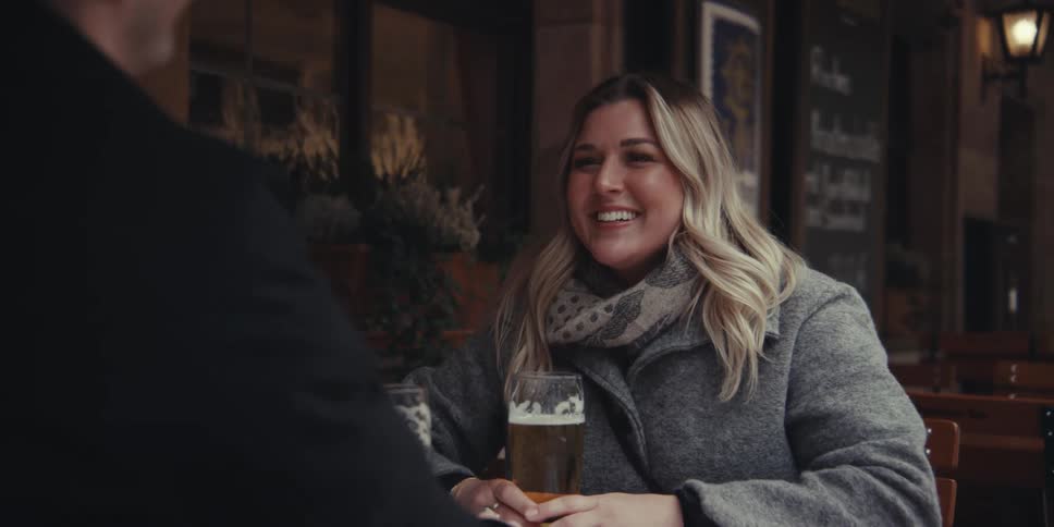 Man and woman sitting at a table as they smile toasting with their beers and holding hands