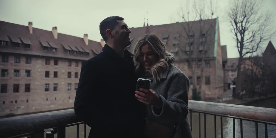 A man and a woman stand on a bridge and take a selfie with a smartphone then they look at the screen laugh and pose together while a building and a river remain visible in the background