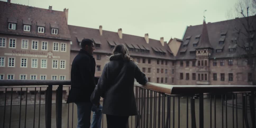 A couple stands close together on a bridge quietly taking in the view of a historic riverside city with old stone buildings in the background