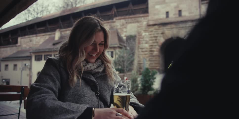 A smiling woman wearing a grey coat and scarf holds a glass of beer while talking with a group of friends at an outdoor table in front of a stone building with an arched gateway