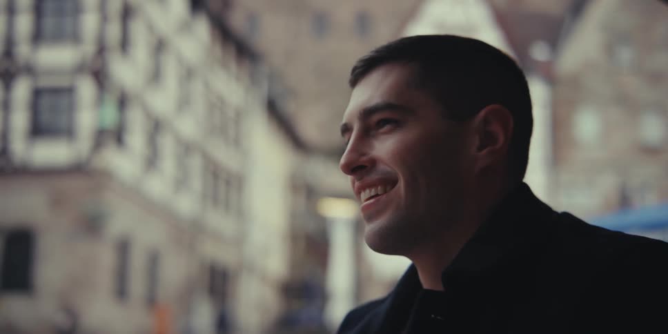 A man wearing a black coat laughs and talks with a woman while holding her hand at an outdoor table in a city with historic buildings