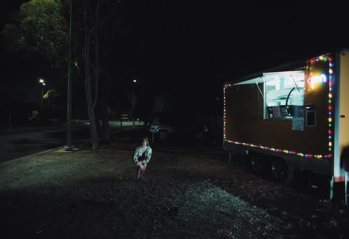 A young woman poses and dances playfully near a brightly lit food truck at night her movement highlighted by the colorful lights against the dark surroundings