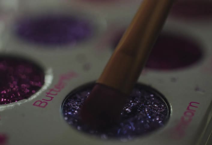 A close view shows a small makeup brush dipping into a circular tray of glittery purple eyeshadow within a palette