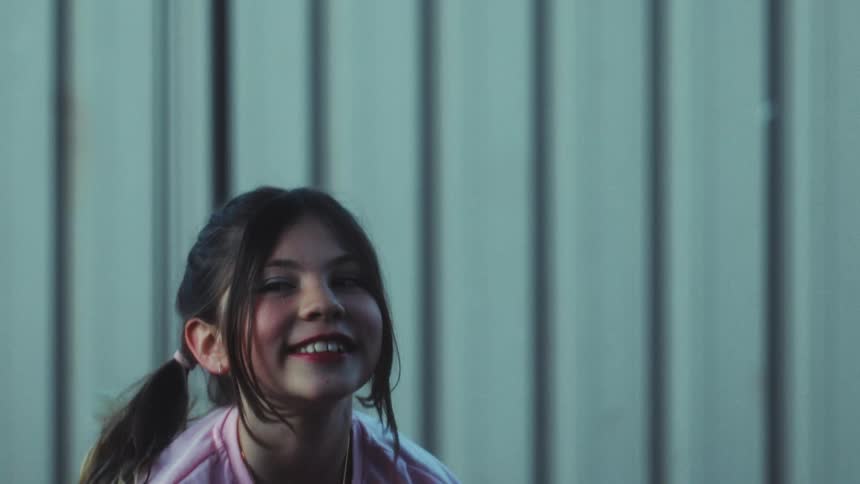 A smiling girl wearing a pink shirt and a gold necklace jumps and dances in front of a grey metal wall