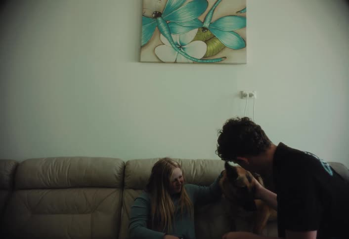 Two people seated on a sofa interact with a dog in a living room