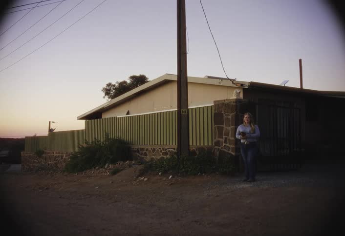 A woman stands by a stone pillar and metal gate outside a house at dusk cradling a small puppy in her arms while looking toward the camera as the evening light fades over the quiet residential street and hills in the distance
