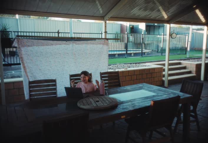Child seated at an outdoor table uses a laptop under a covered area